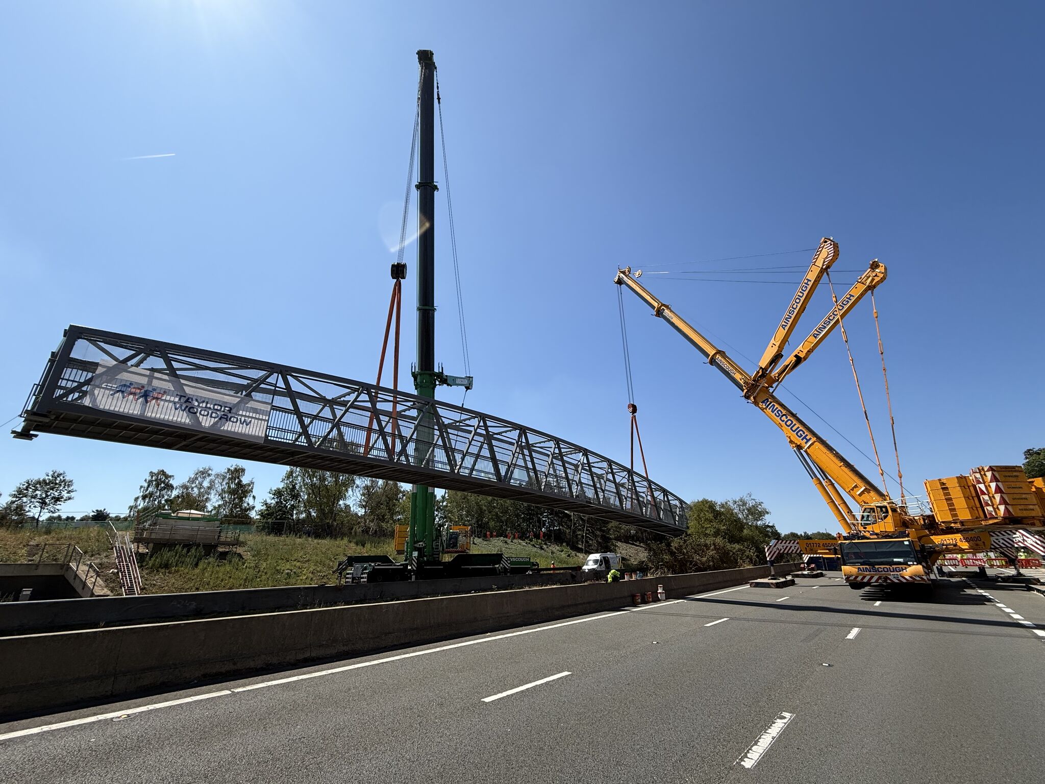 Two cranes lifting a steel footbridge over a motorway